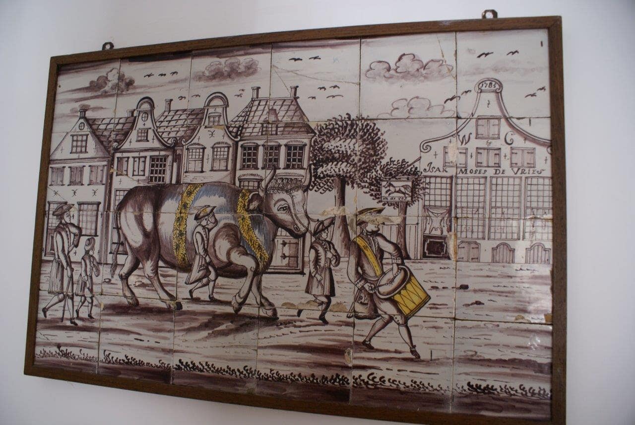 Het tegeltableau met rechts de slagerij. In 1785 opende Izak Moses de Vries een koosjere slagerij in de Zwolse Diezerstraat. Hij is een voorouder van Fien in de zesde generatie. Het slagersvak was één van de weinige ambachtelijke beroepen die Joden vóór de burgerlijke gelijkstelling in 1796 mochten uitoefenen. Bron: www.geschiedenisjoodszwolle.nl/marcus/. Beeld: Judith Marcus.
